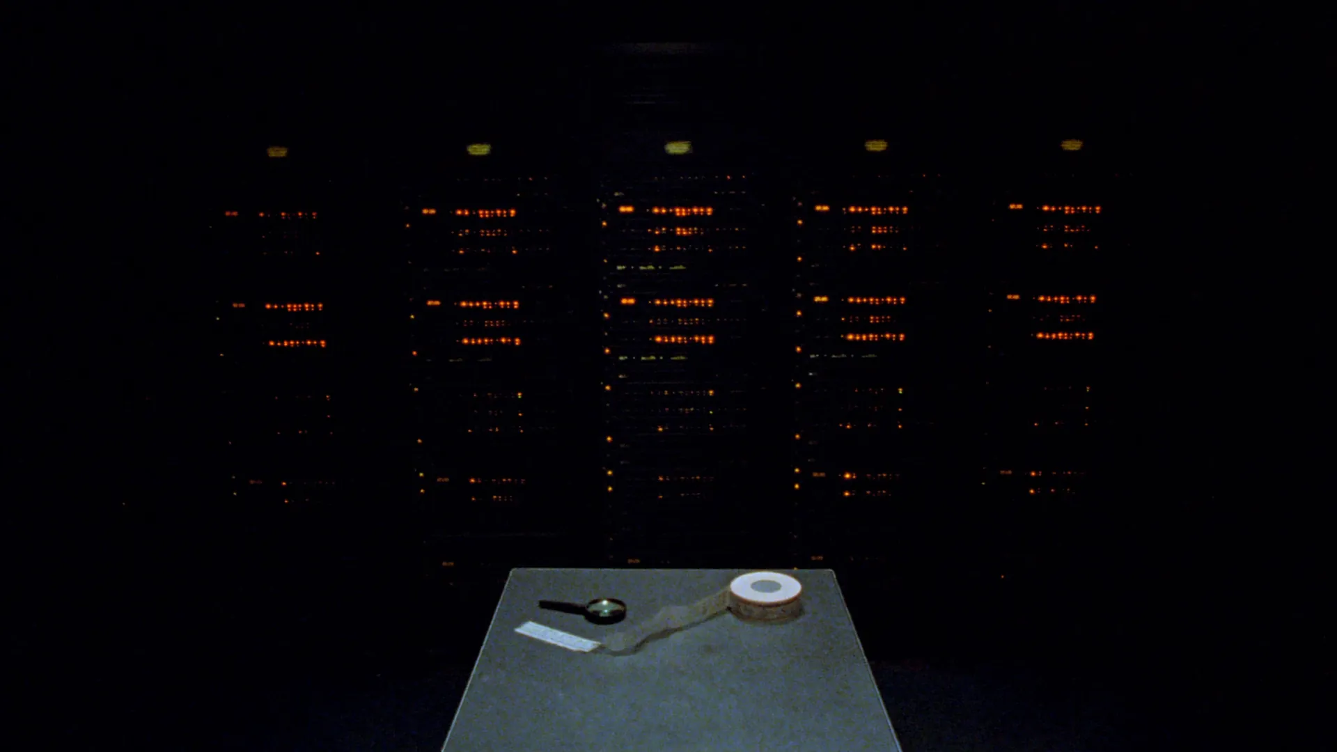 A dark data room with racks of amber status lights and a reel of ticker tape on a steel table